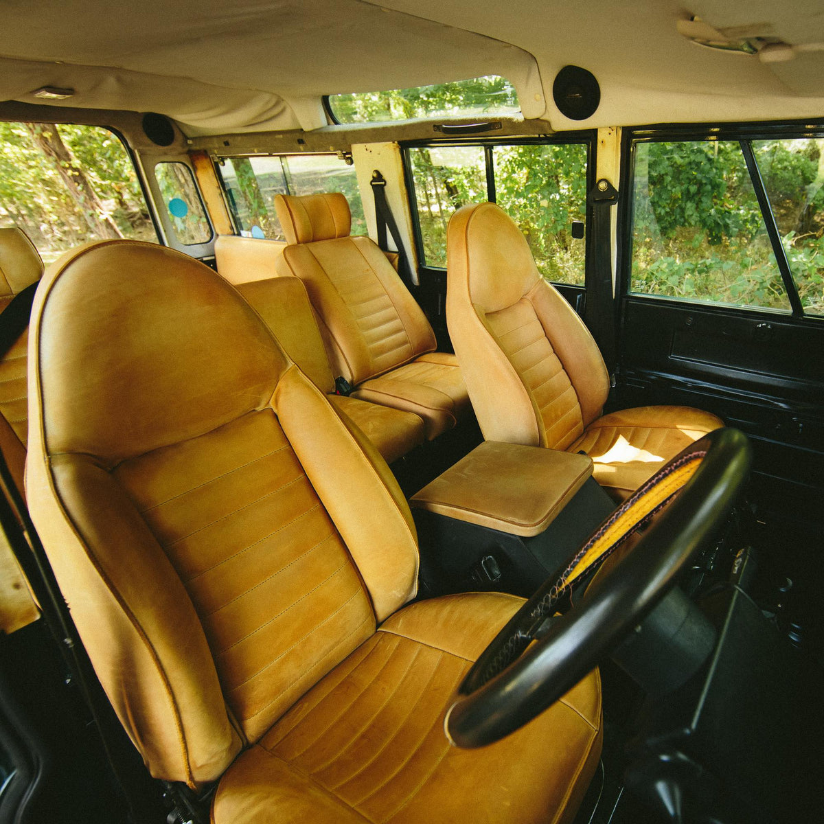 Inside a vintage four-by-four: sun-faded tan leather seats front and rear, a leather centre armrest and a steering wheel, with trees outside the open windows.