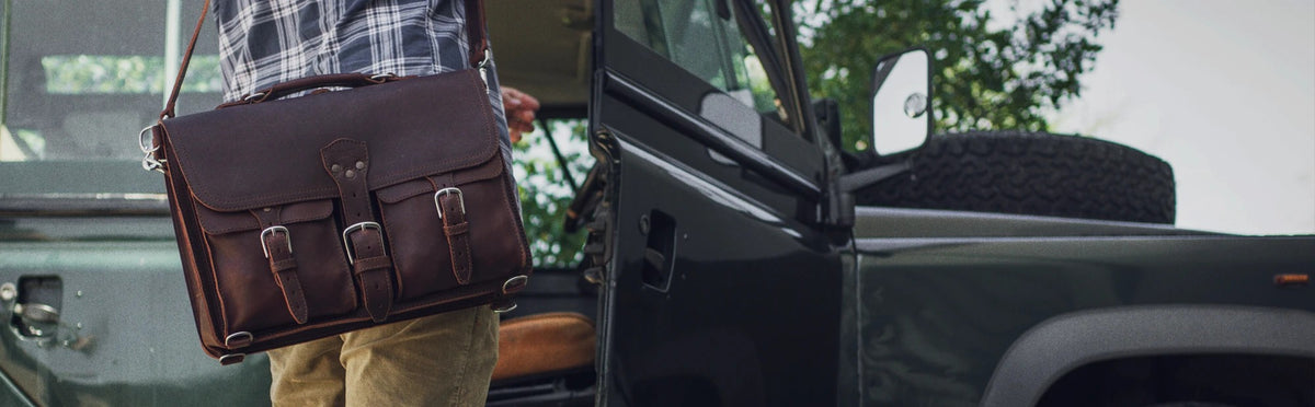 Thin Front Pocket Leather Briefcase in Dark Coffee Brown carried by the handle by a person in a plaid shirt beside a dark truck