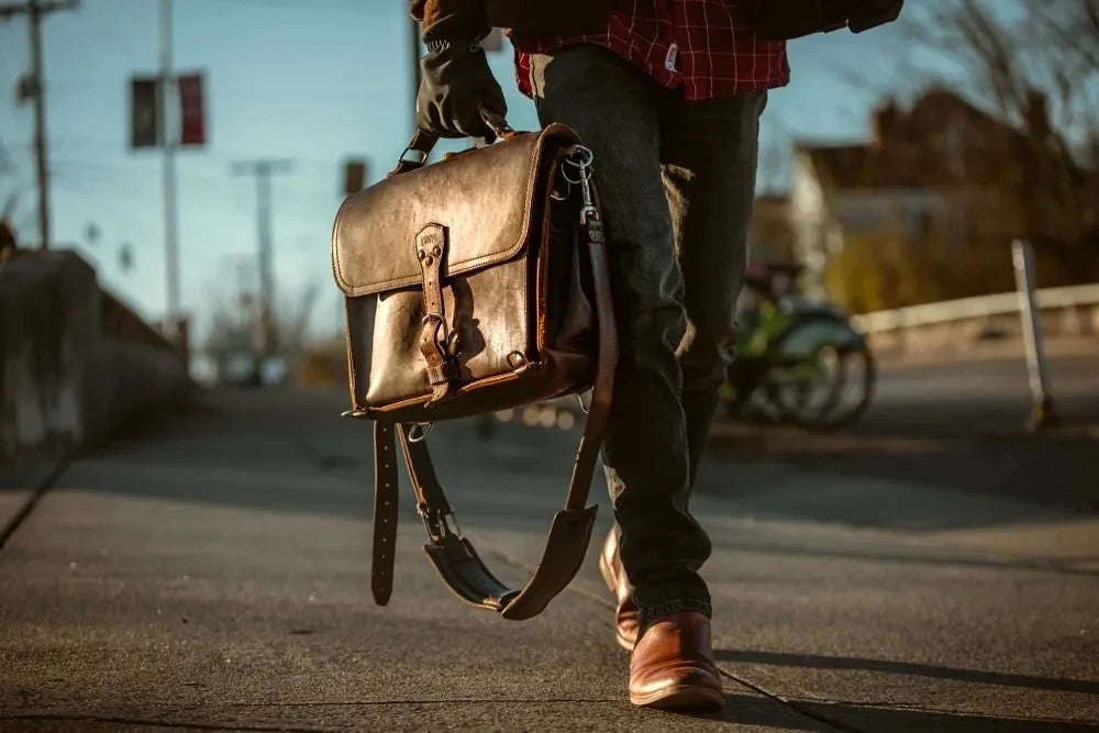 Man in a plaid shirt carrying a dark brown full grain leather slim briefcase down a sunlit street