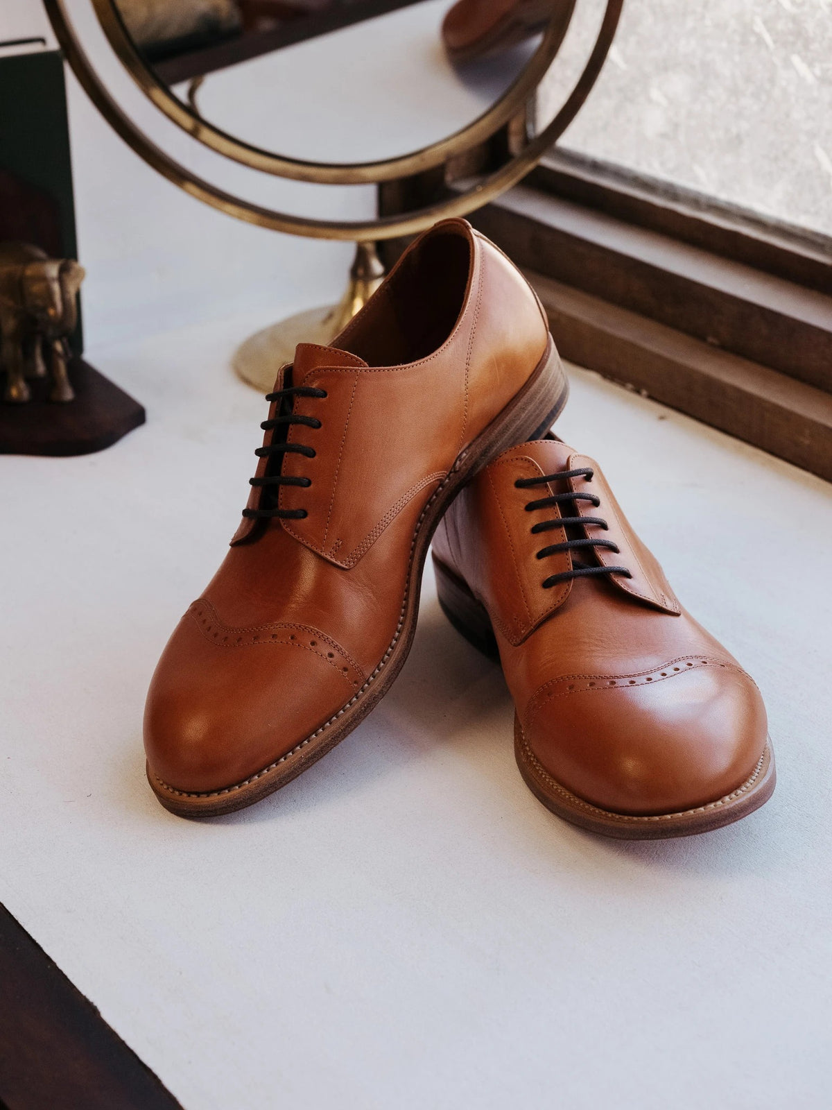 Toe Cap Shoes - English Tan, a pair stacked on a sunny windowsill beside a brass mirror