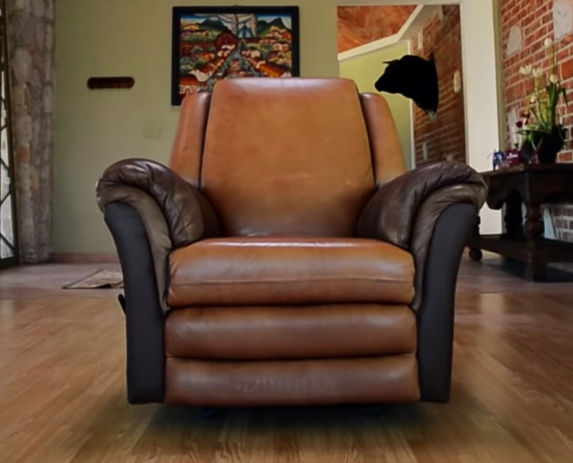 A tan leather recliner with darker brown arms on a wooden floor, in a living room with a painting, a mounted bull's head and brick walls behind it.