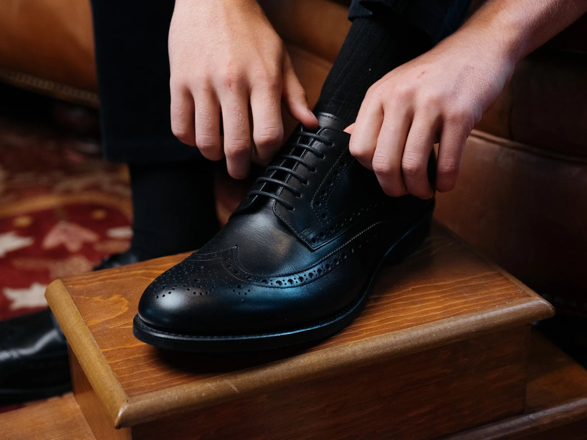 Wingtip Shoes - Raven Black on foot, hands tying the laces with one foot up on a wooden step