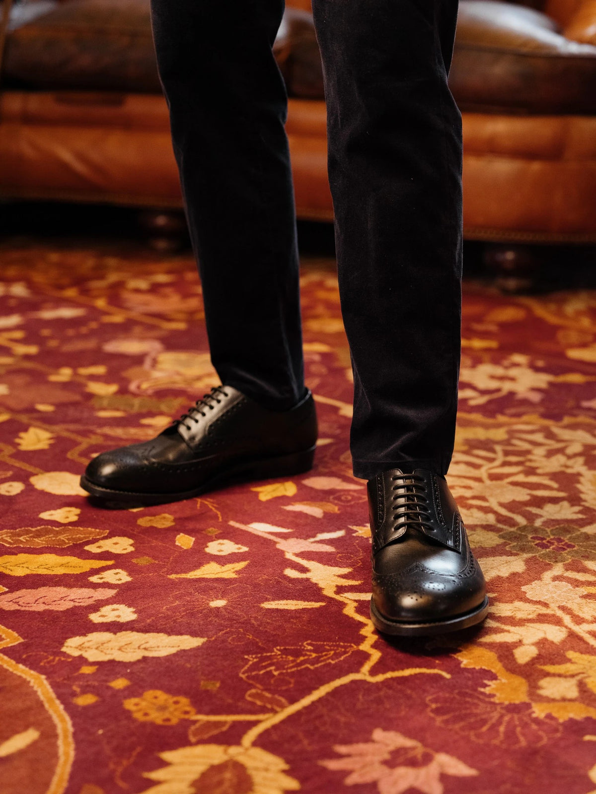 Wingtip Shoes - Raven Black on foot, a person in black trousers standing on a red patterned rug