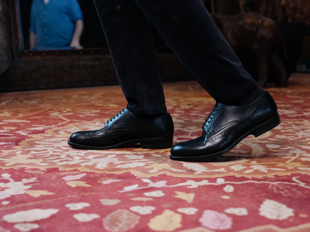 Wingtip Shoes - Raven Black on foot, side view mid-stride in black trousers across a pink patterned rug