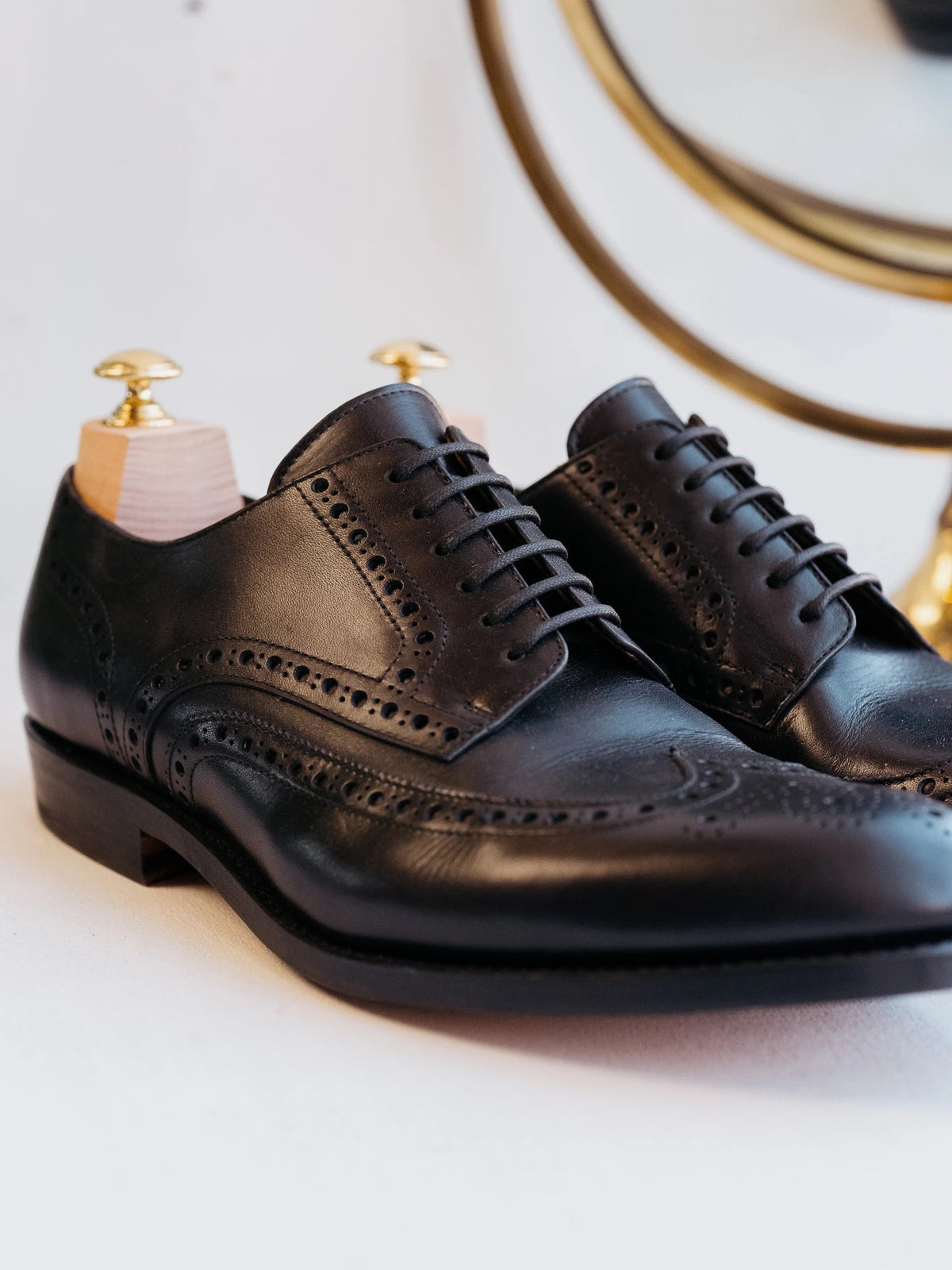 Wingtip Shoes - Raven Black, three-quarter view of a pair with wooden shoe trees beside a brass mirror
