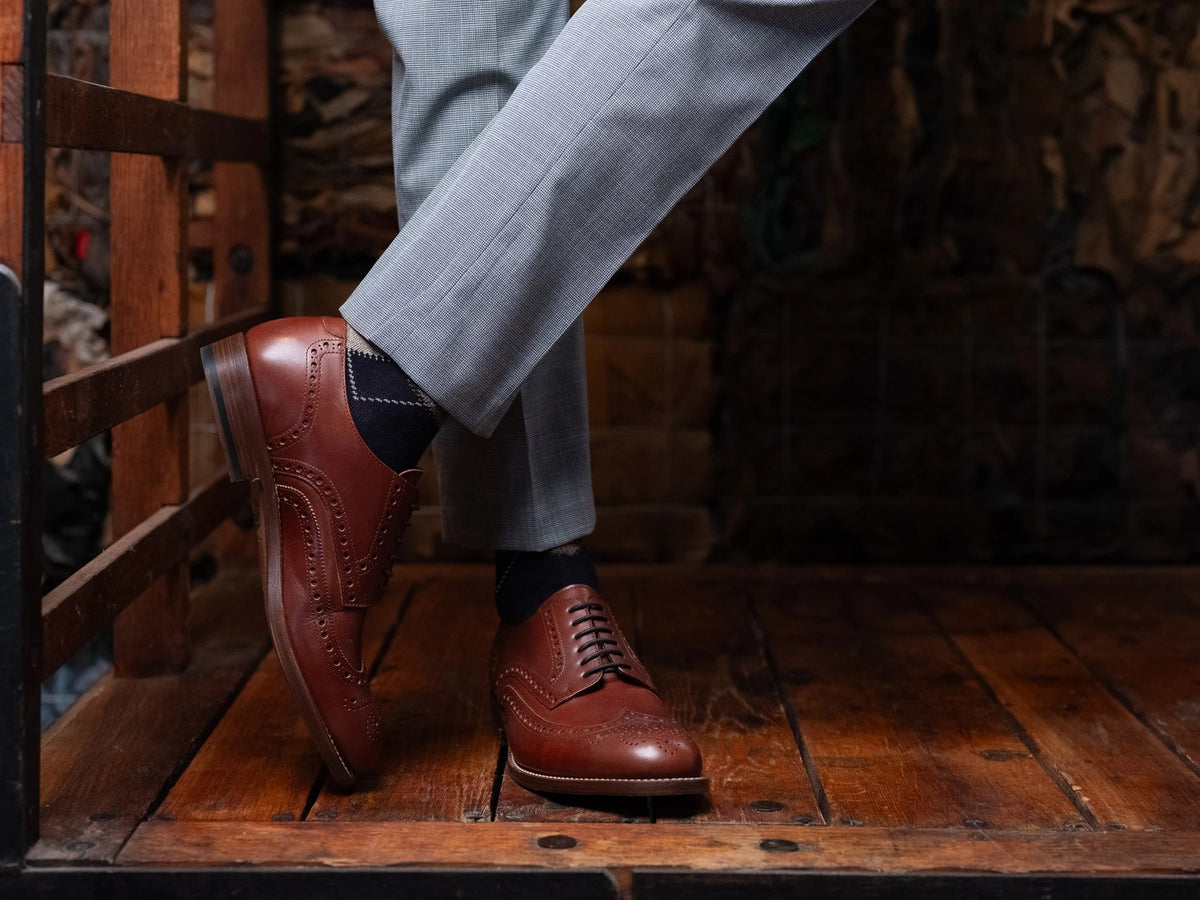 Wingtip Shoes - Chestnut worn with crossed legs and grey trousers on a rustic wood platform