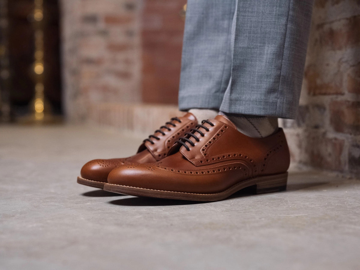 Wingtip Shoes - Natural Tan on foot, side view of a pair on a concrete floor with grey trousers and brick wall